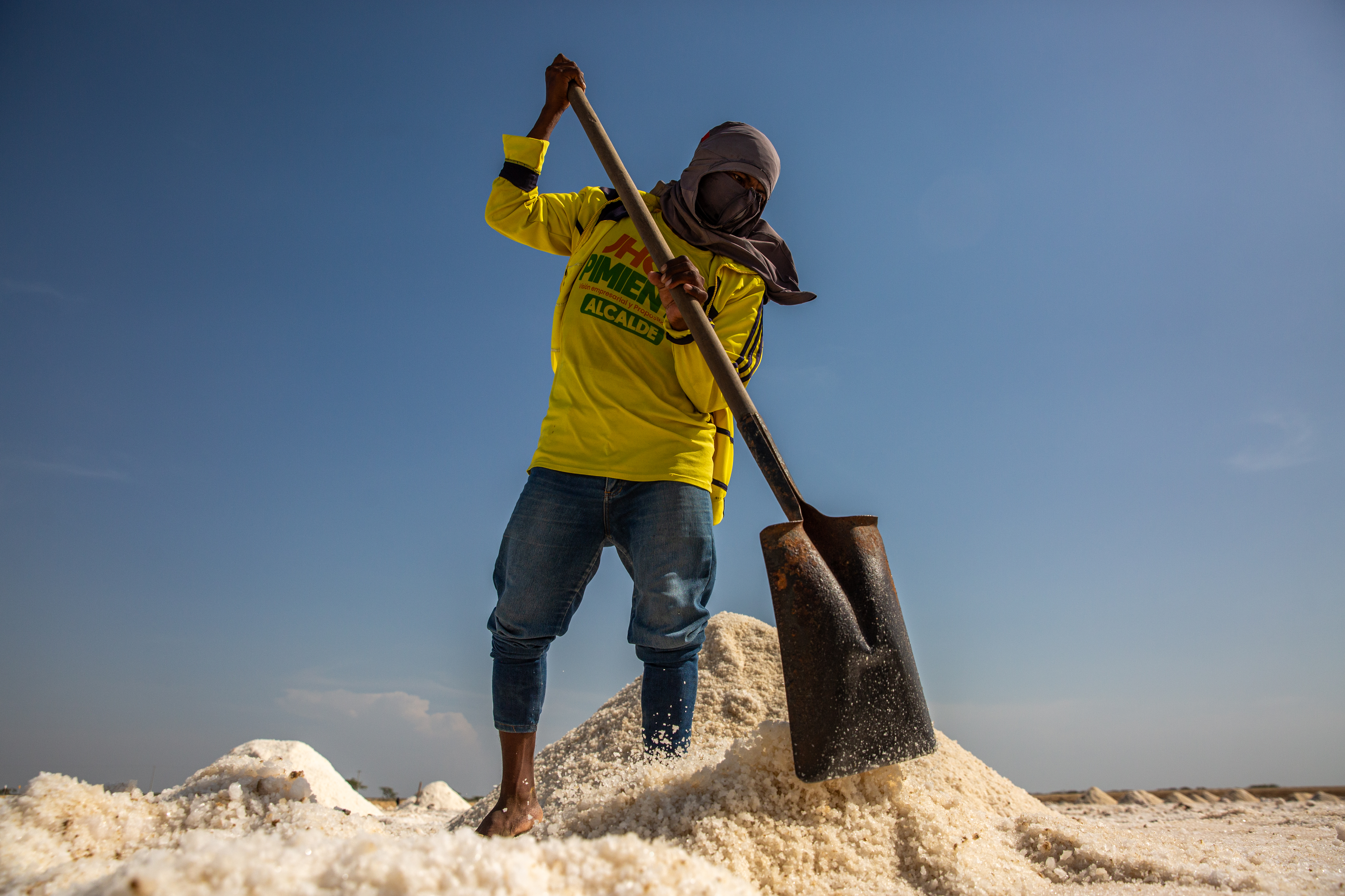 A man — his head wrapped in a scarf to protect against the wind and heat — uses a shovel to pile up salt.