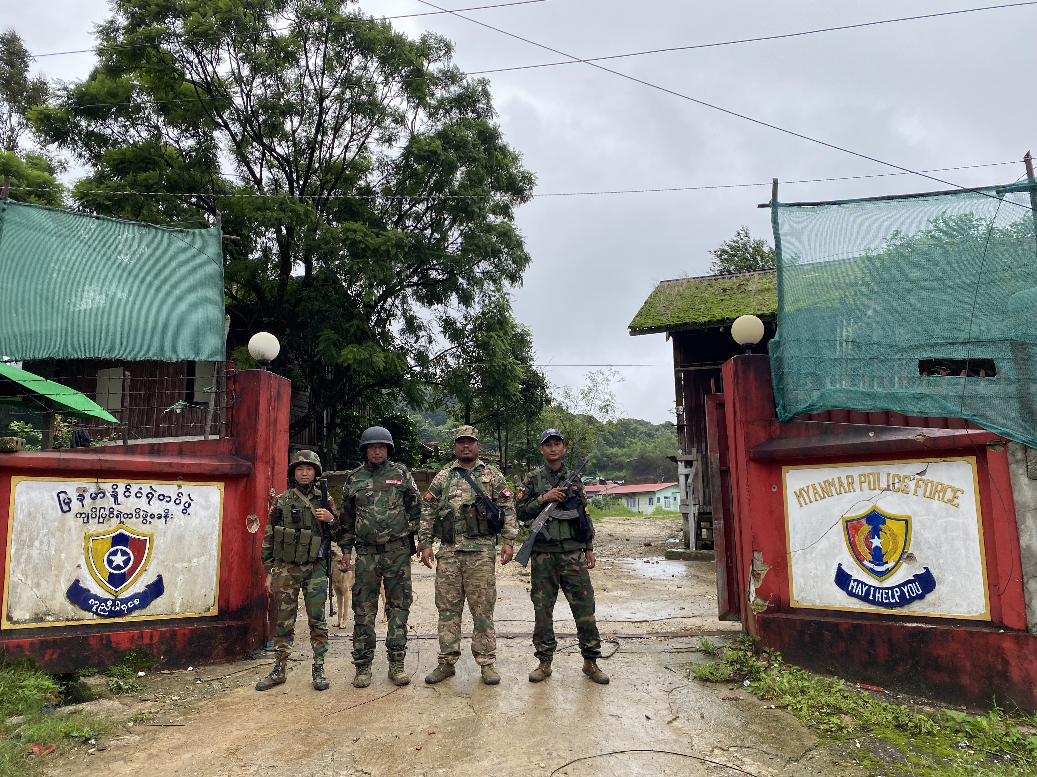 Anti-coup fighters standing at the entrance to a Myanmar police station. The gates are open. It looks like it's been raining.