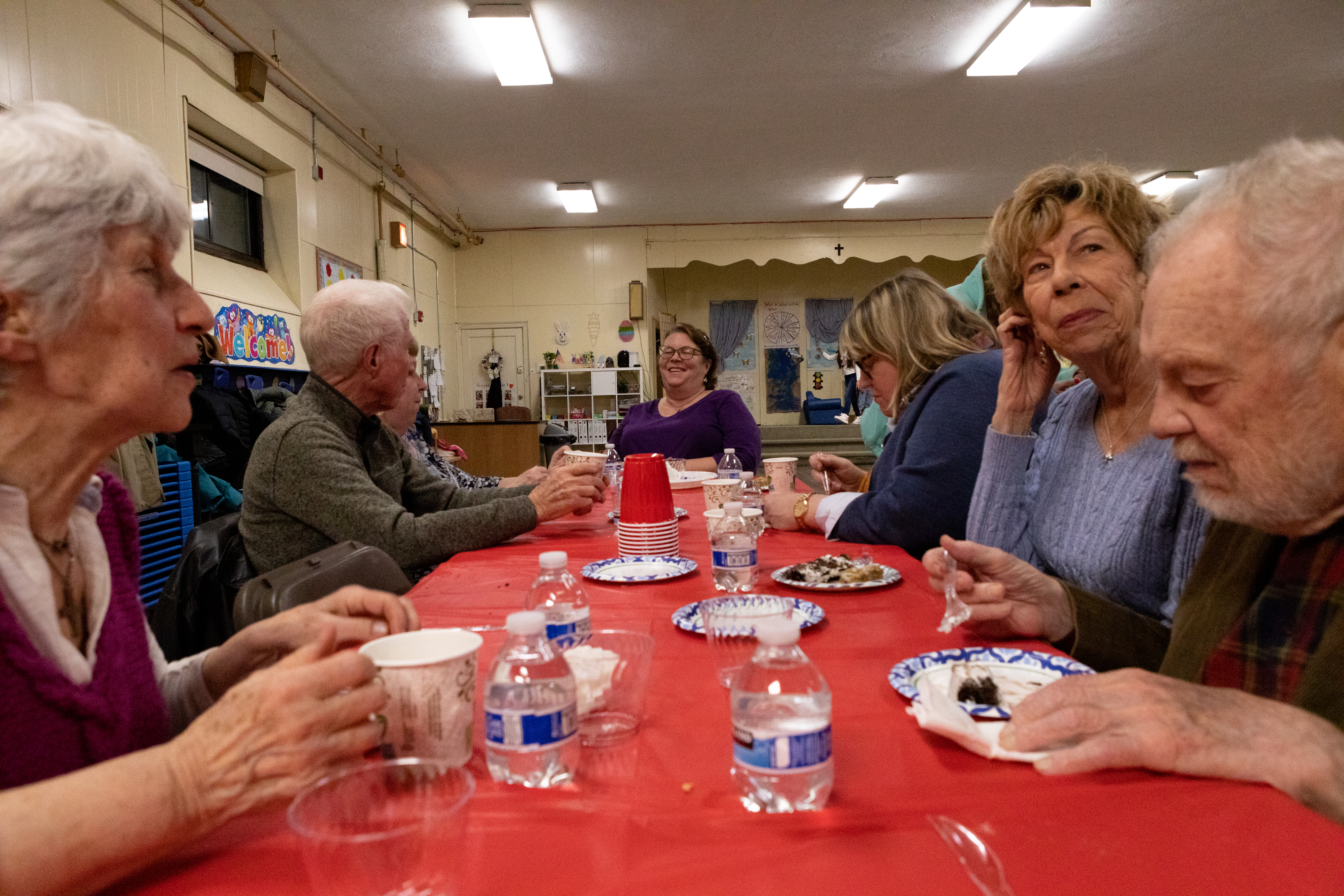 Parishioners gather around a long table with a red table cloth to eat cake off of paper plates.