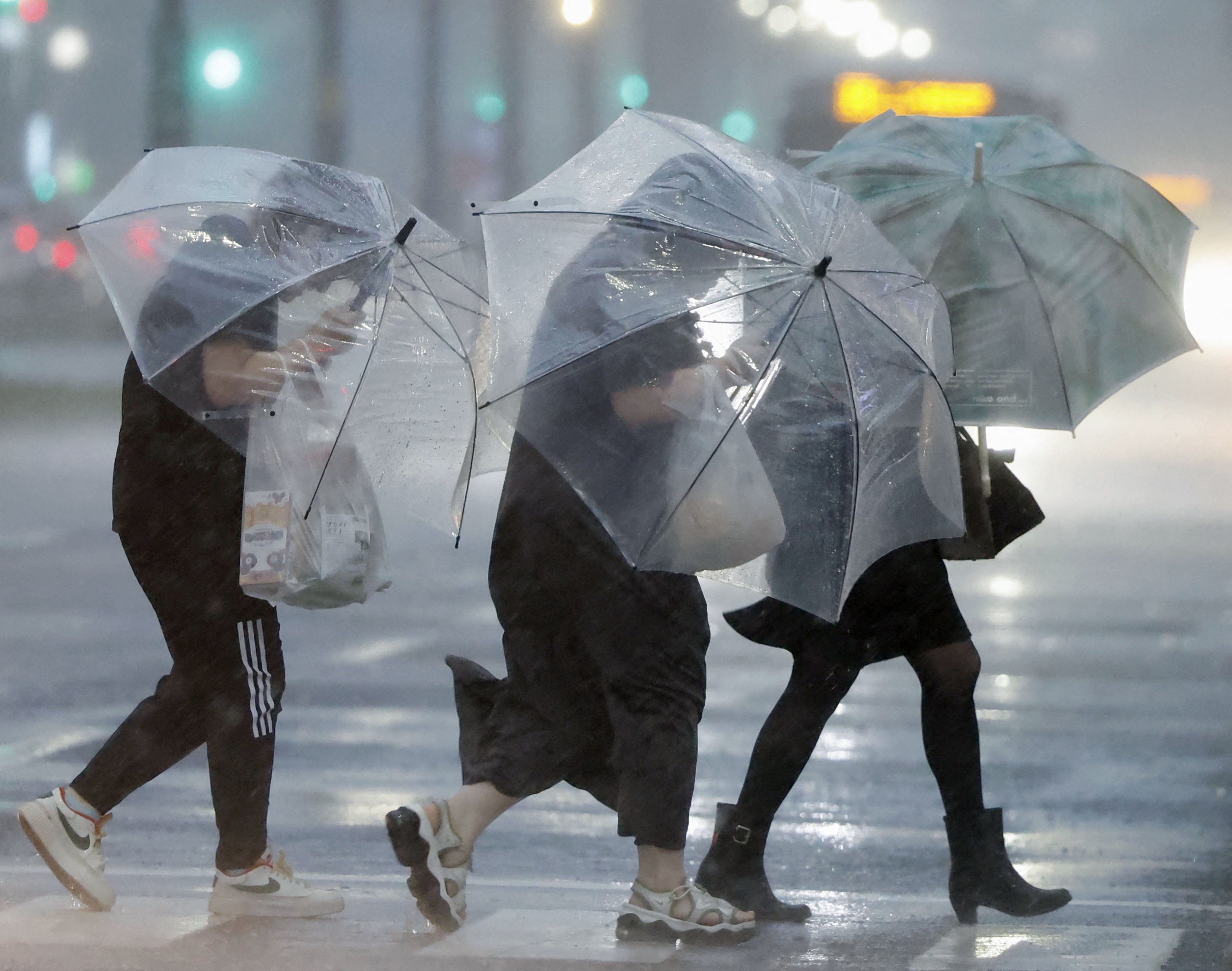 Three people struggling under umbrellas in Kagoshima. The pavement is really wet. The rain is extremely heavy. The wind is twisting their umbrellas.