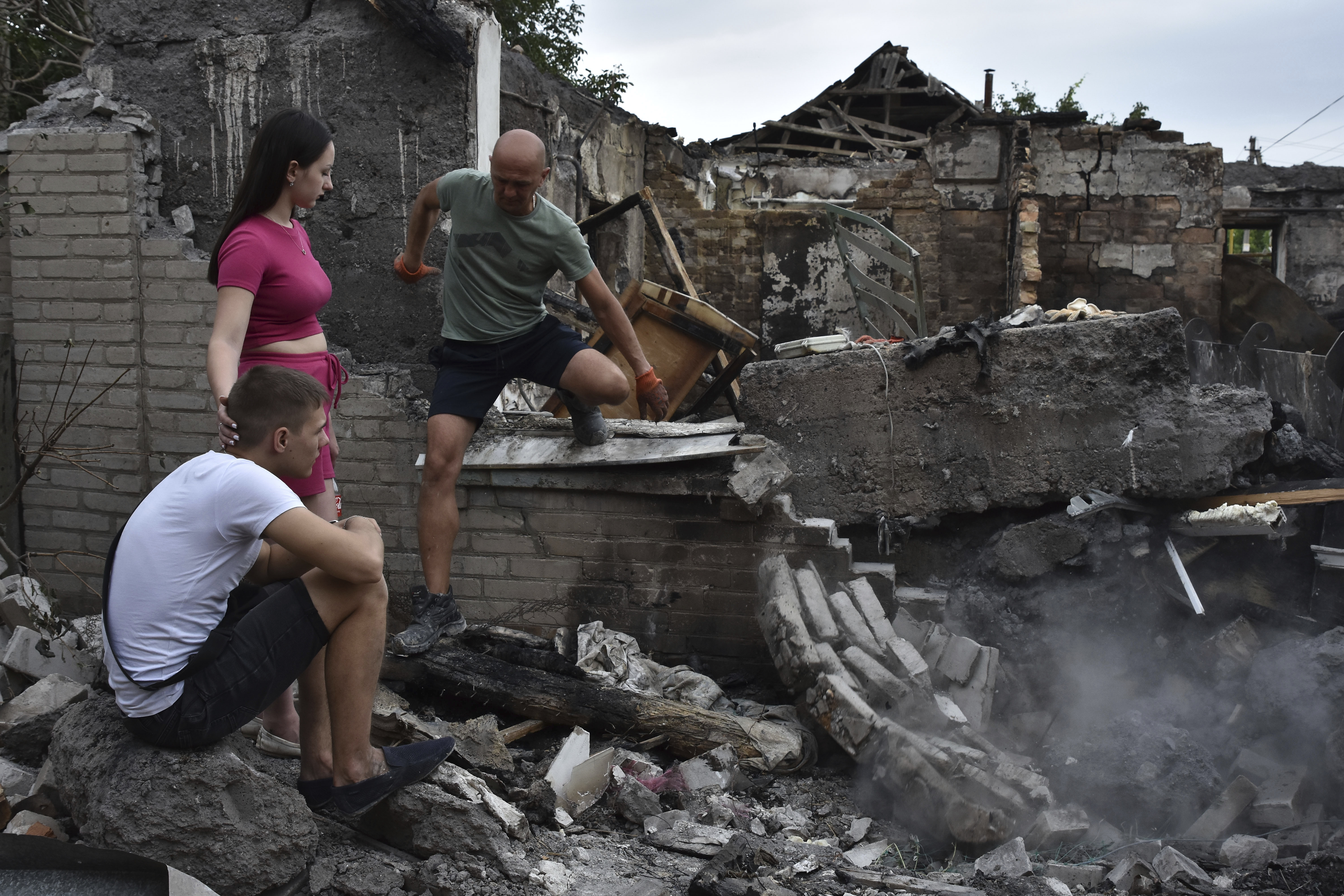 A couple amid the rubble in front of their ruined house. The woman is standing with her hand on her partner's shoulder. He is sitting in the rubble and looks shocked. A third man is climbing over one of the ruined walls.
