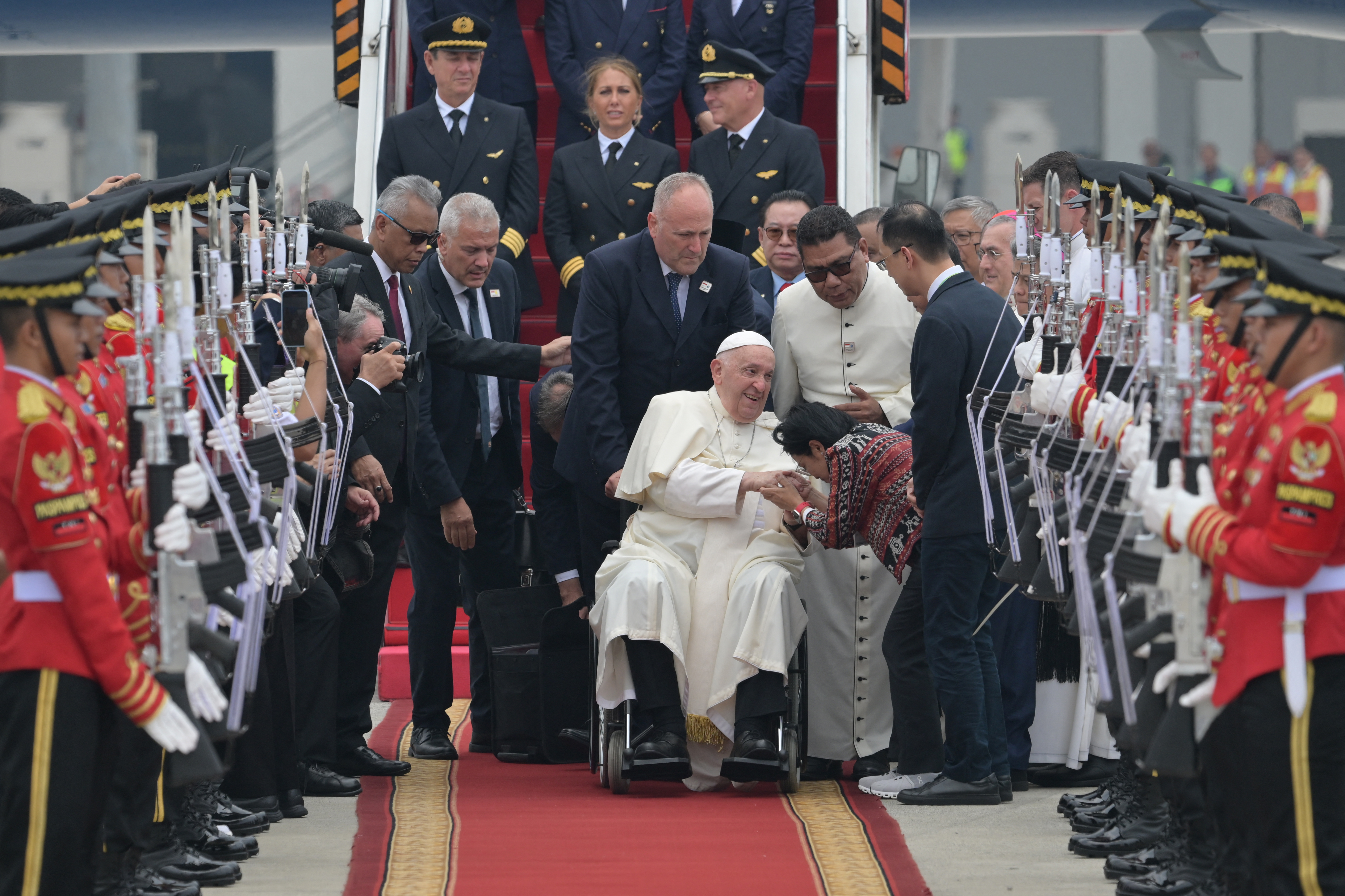 The pope being welcomed at Jakarta airport. He's in a wheelchair. A wellwisher is bent towards him holding his hand. He is smiling. There is an honour guard and various men and officials behind him. The plane's pilot and crew are also o the steps behind him.