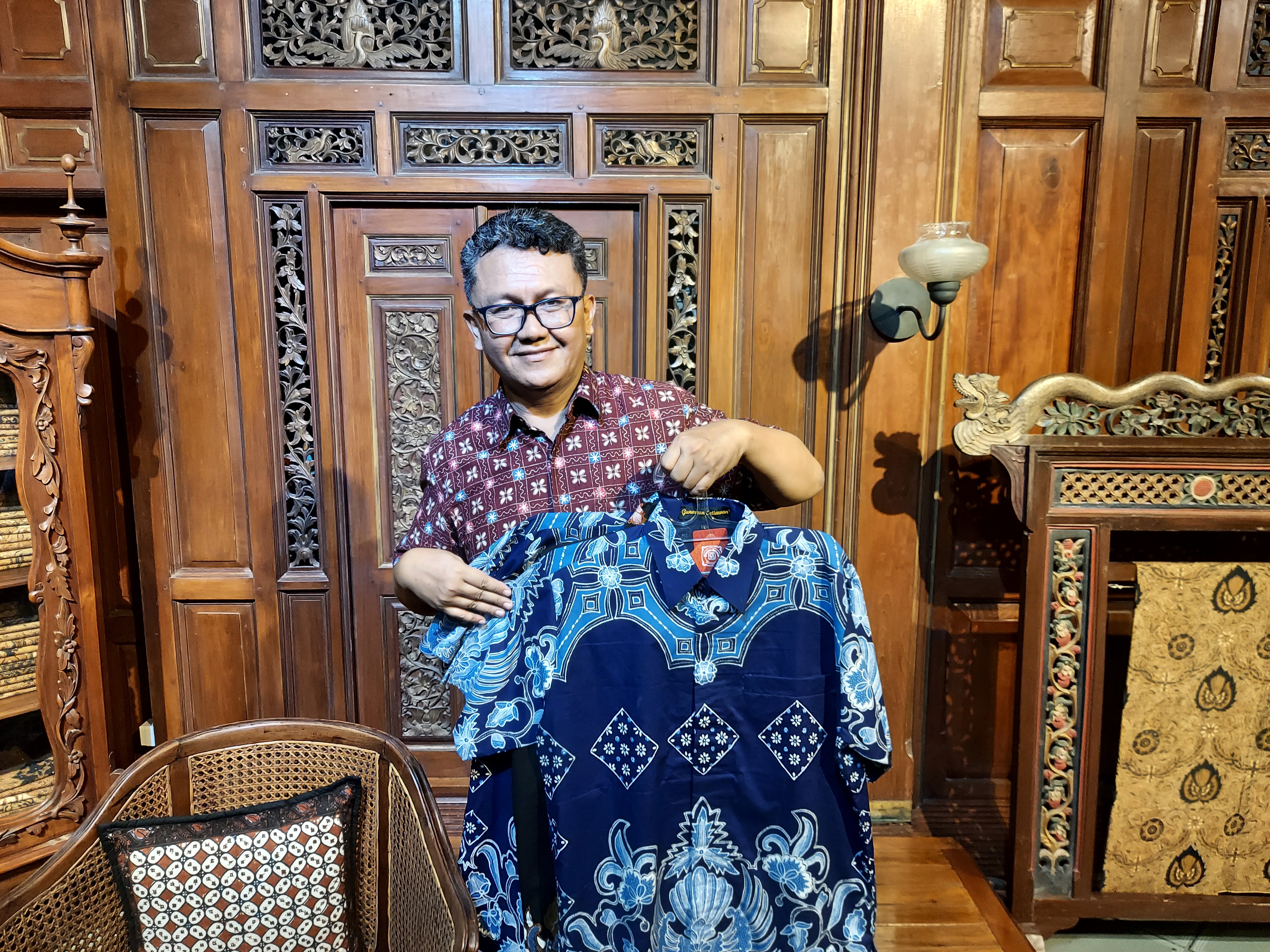 Gunawan Setiawan. He is wearing a marron-coloured batik short and holding up shirts with blue designs, He is in a wood-pannelled room with rattan-sated chairs. 