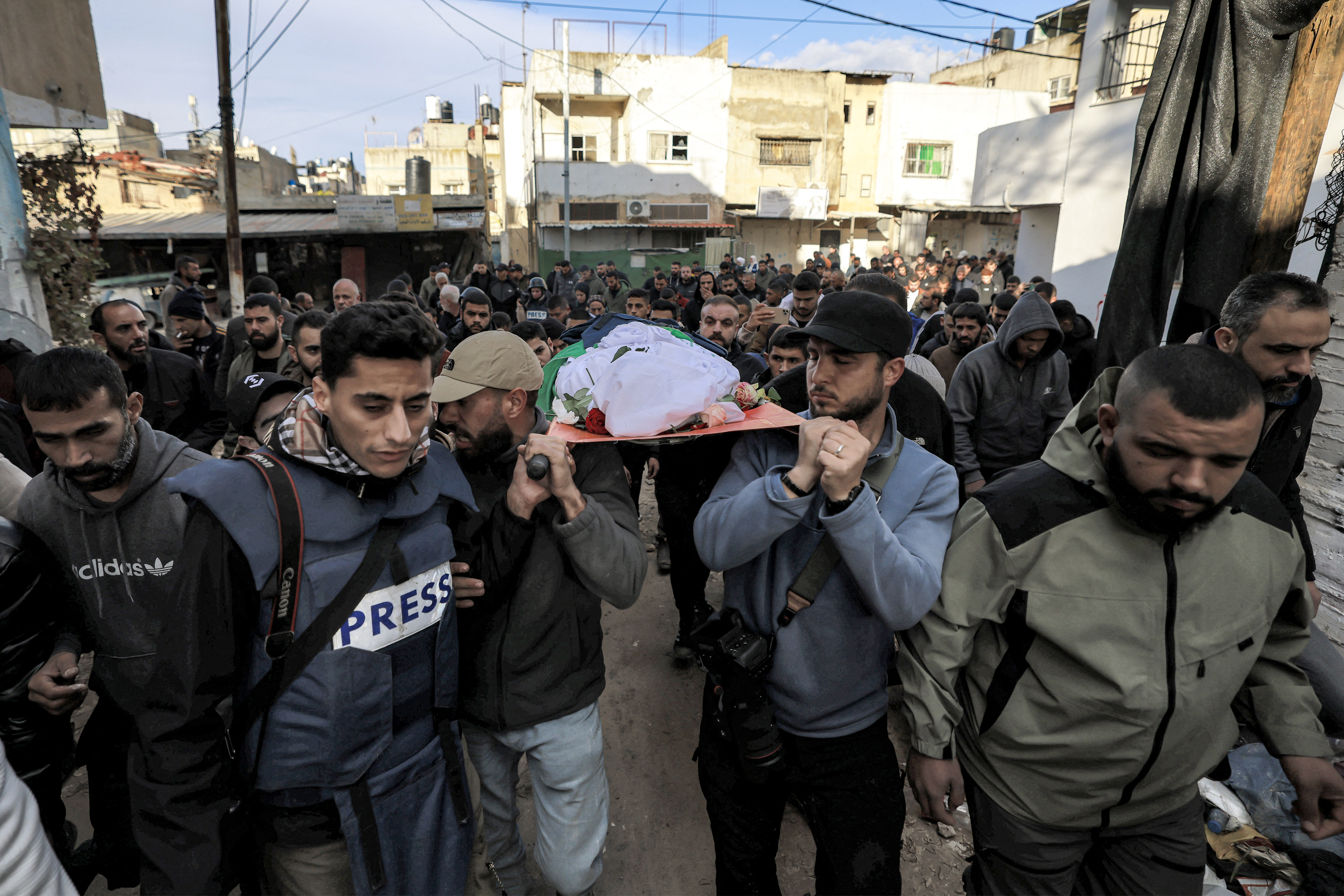 people including a man wearing a press vest carry a body on their shoulders through the street