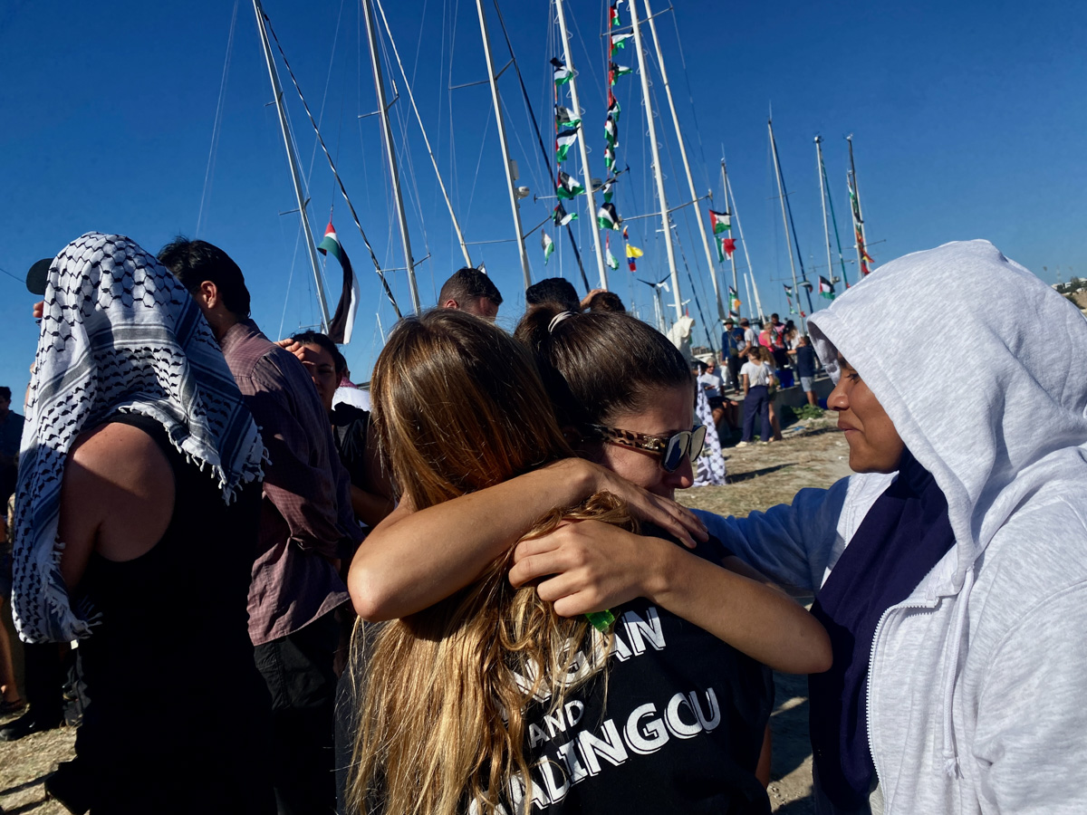 Female volunteers hugging their goodbyes with boats in the background, their masts full of fluttering Palestinian flags