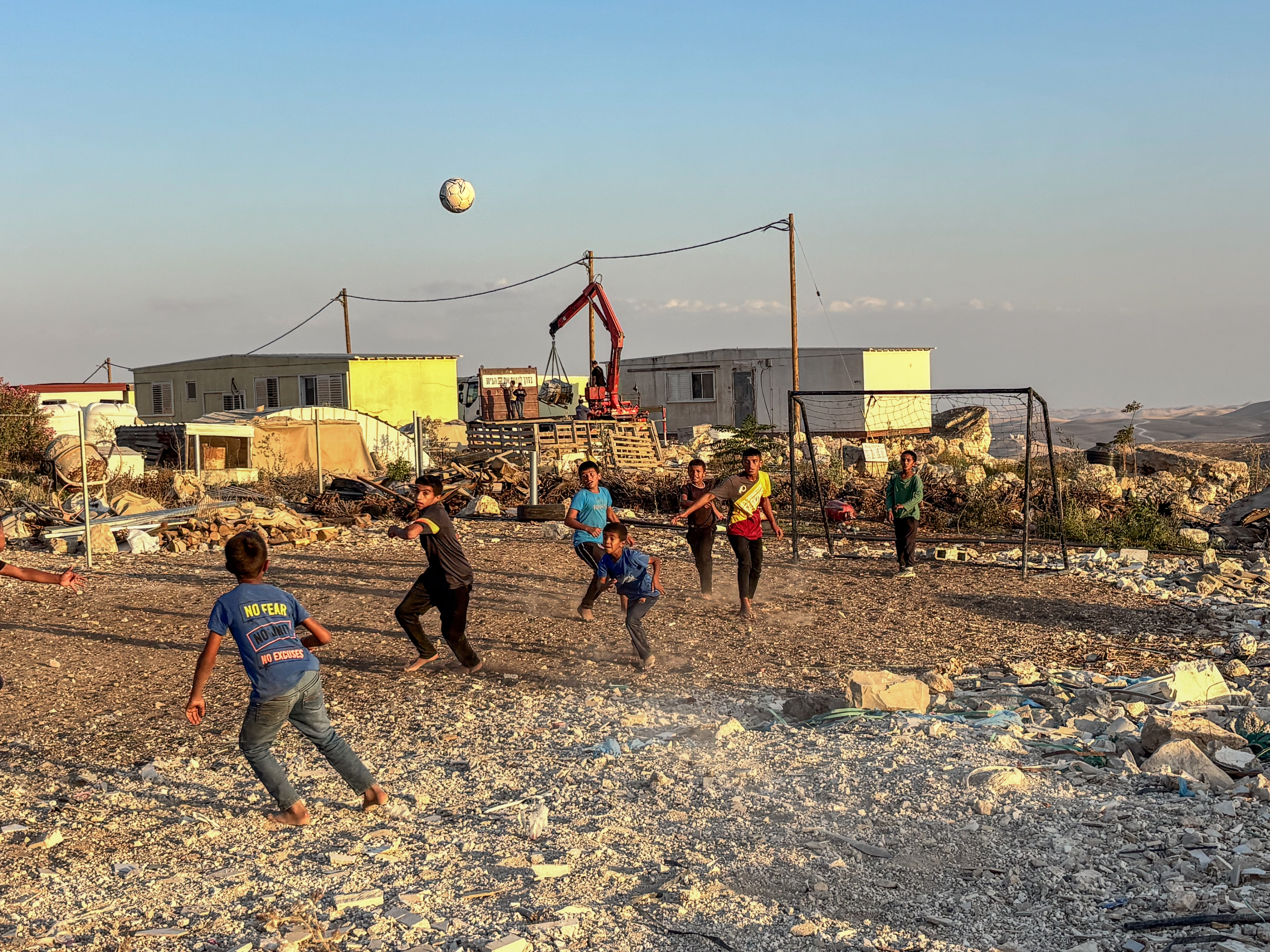 Children in street clothes playing football in their bare feet on a dirt pitch, engrossed in their game. Squatting threateningly behind them are the caravans set up by Israeli settlers to hem in the village