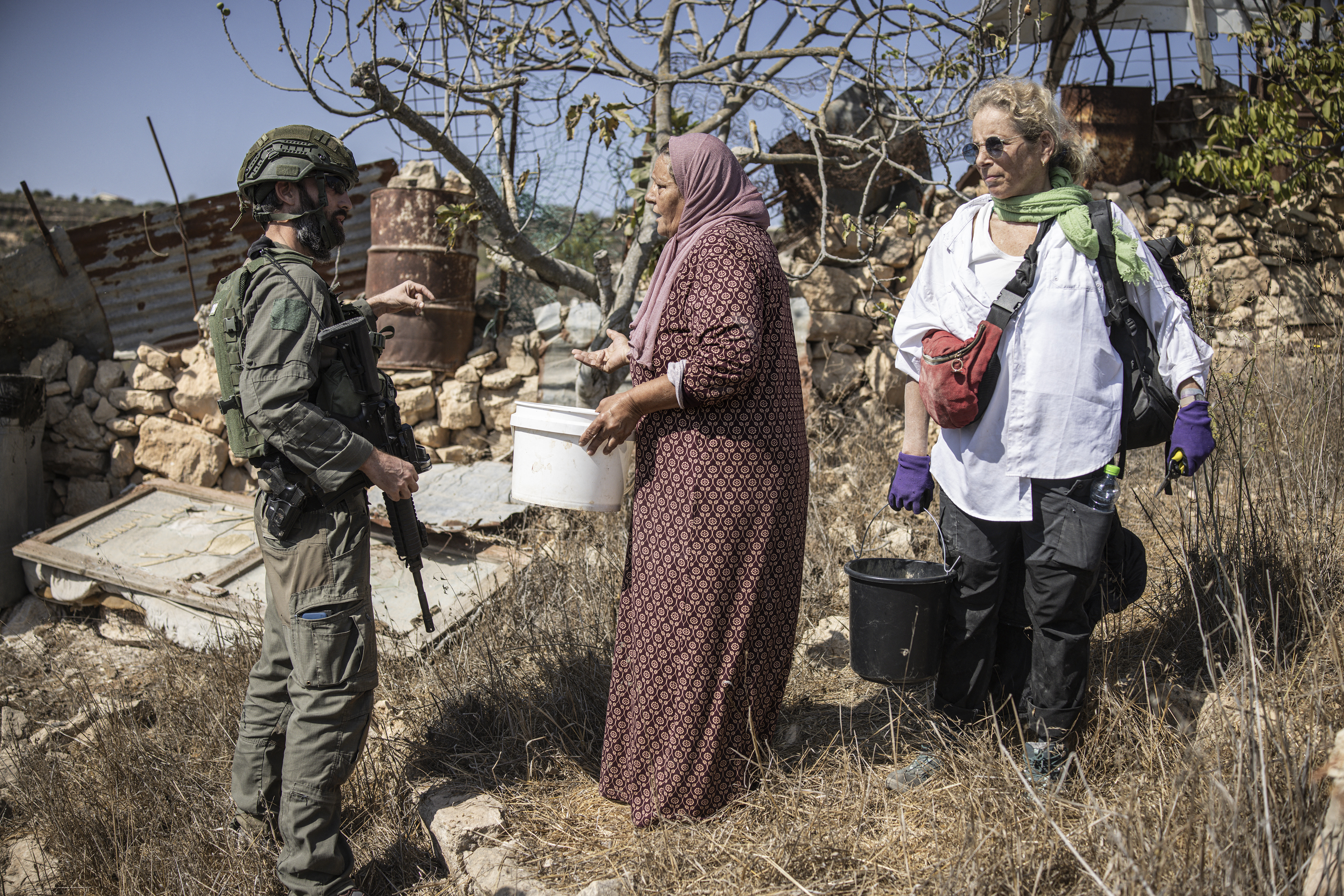 a lady in a pink dress and head scarf holds a white bucket and faces an armed soldier