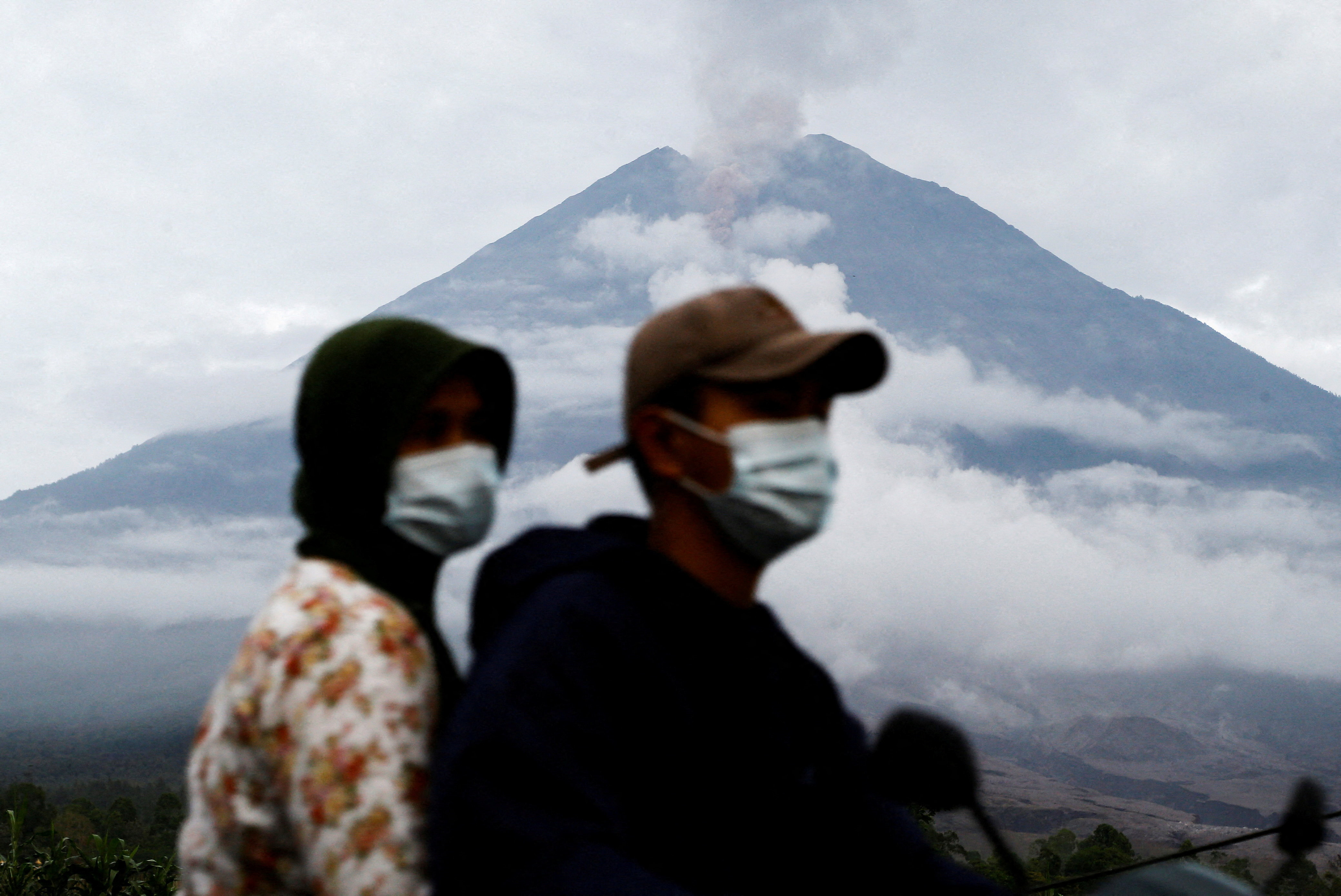 Locals ride a motorbike as Mount Semeru spews volcanic ash in the background, in Supiturang village in Lumajang, East Java province, Indonesia, November 20, 2025