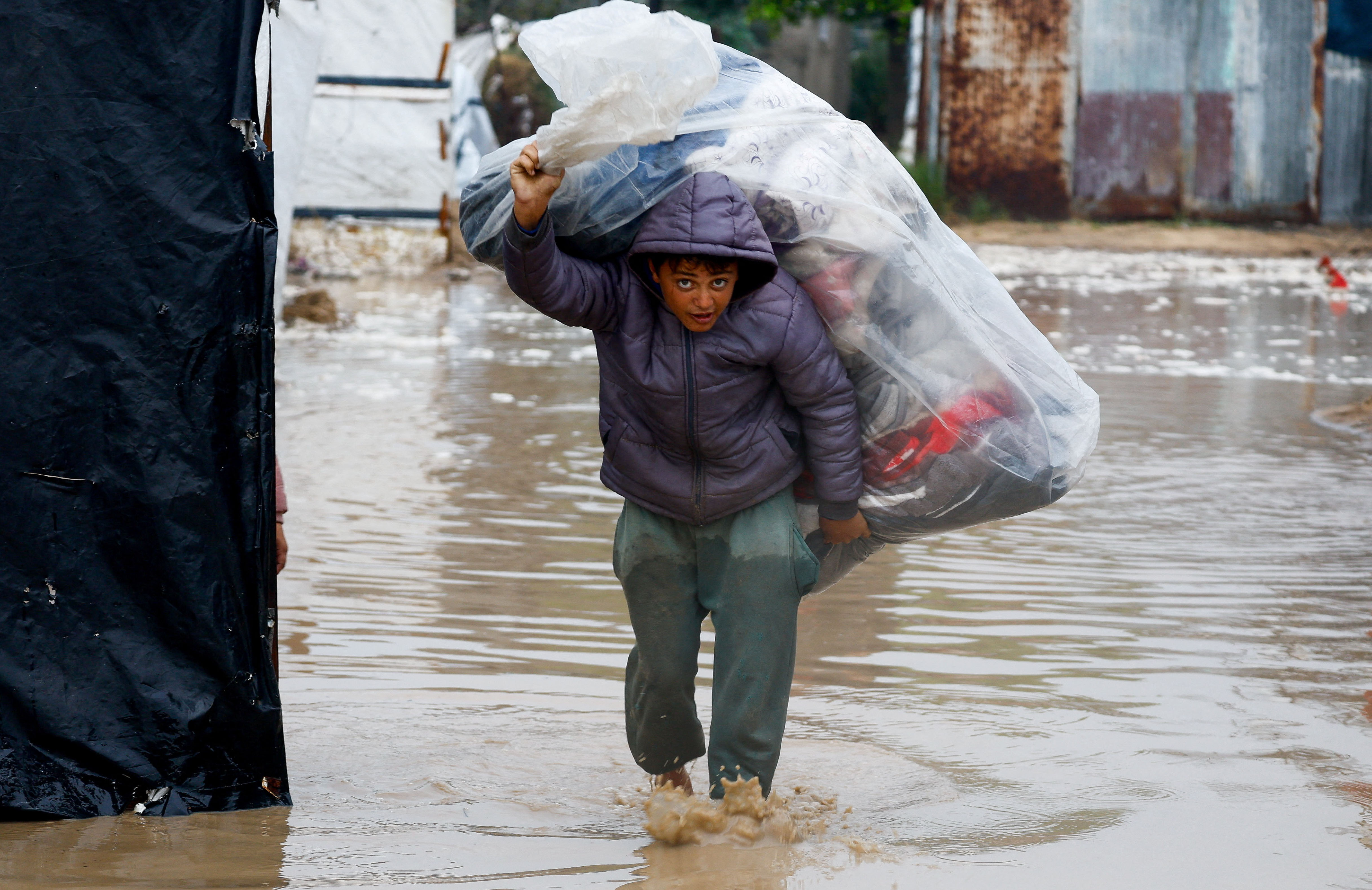 A displaced Palestinian carries belongings in a flooded tent camp on a rainy day in Nuseirat, central Gaza Strip, December 12, 2025. [Mahmoud Issa/Reuters]