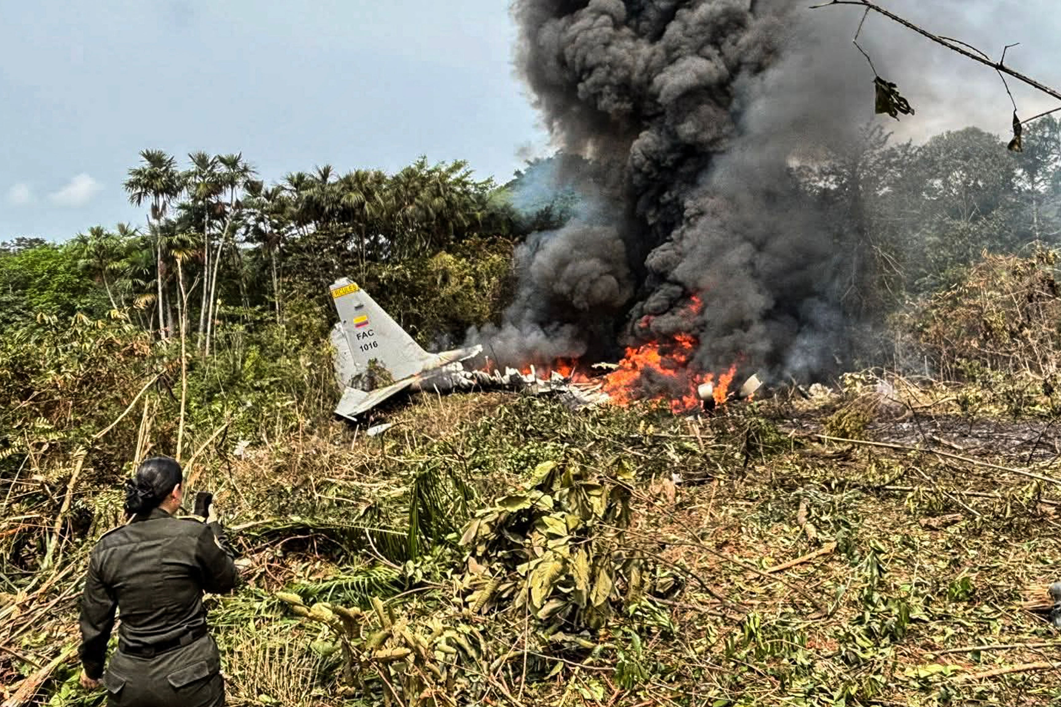 Flames and thick black smoke rise from an Air Force Hercules that crashed during takeoff, as a member of the Colombian Police stands nearby, in Puerto Leguizamo, Colombia, near the southern border with Ecuador, on March 23, 2026. [Daniel Ortiz/AFP]