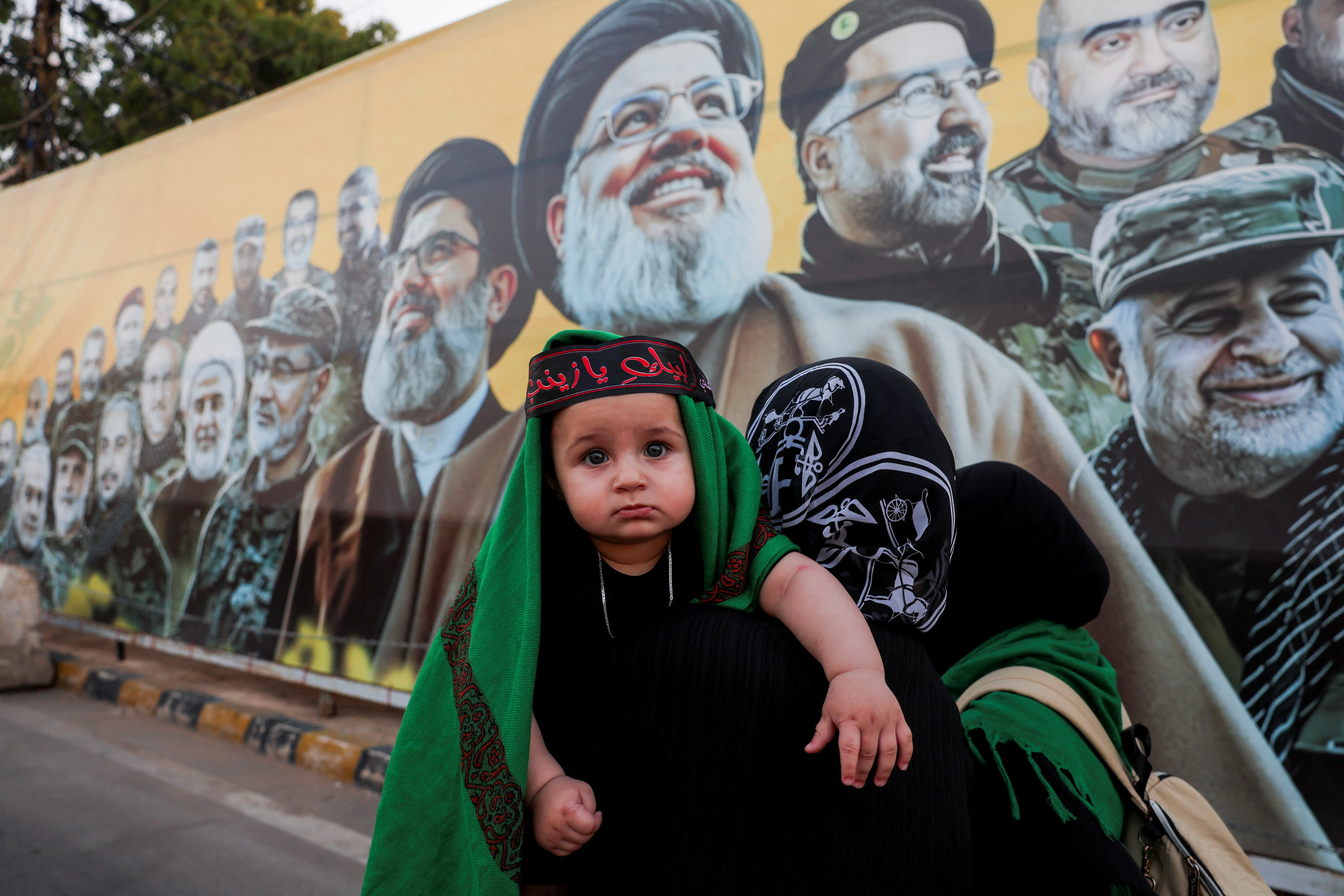 Women carrying a child walk past a mural depicting former Hezbollah leader Hassan Nasrallah and other senior figures, on the day Hezbollah chief Naim Qassem delivers a televised speech, near the burial site of Hassan Nasrallah on the outskirts of Beirut, Lebanon, June 17, 2026