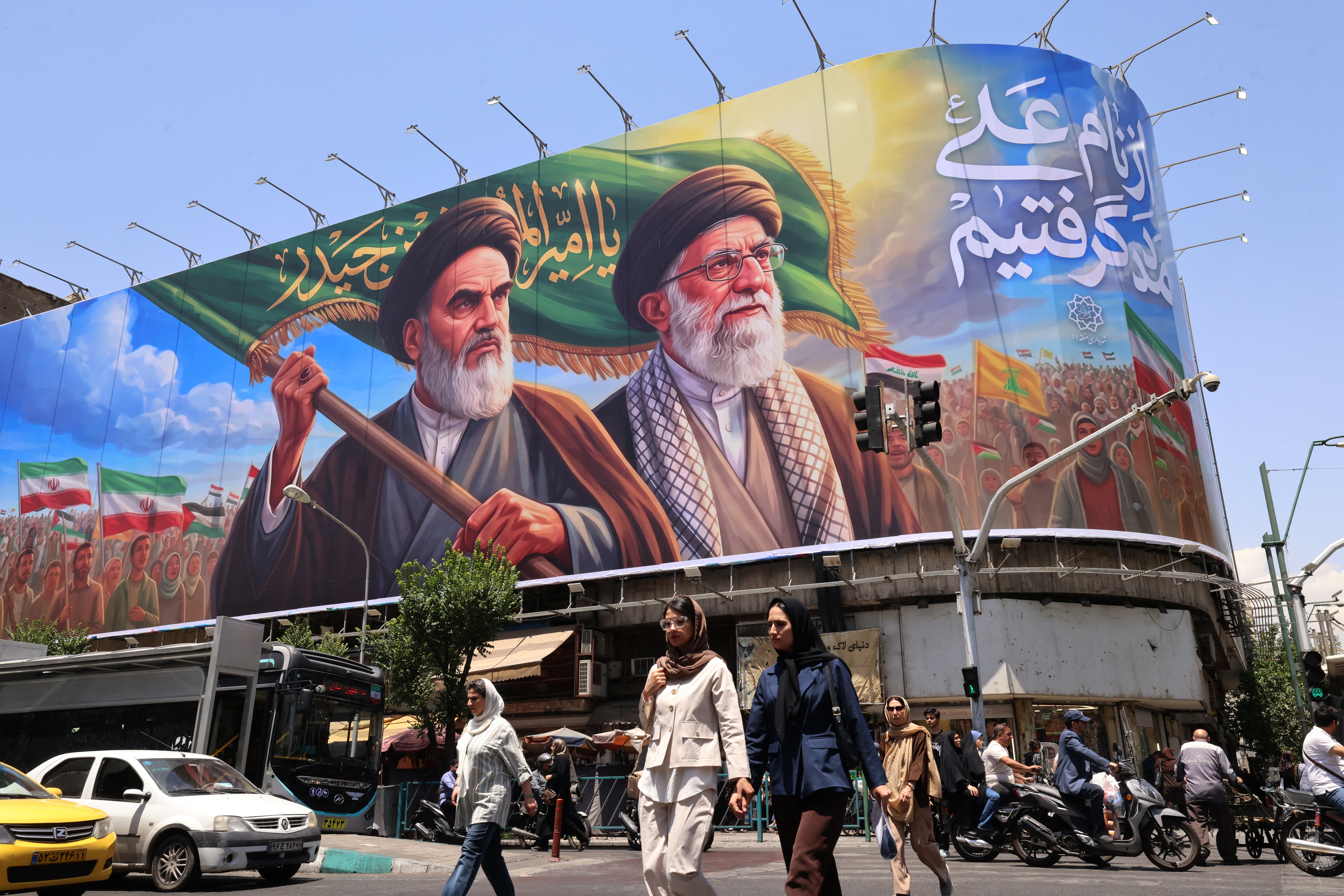 TOPSHOT - People cross the street past a large billboard showing portraits of Iran's late supreme leader, Ayatollah Ruhollah Khomeini (L) and slain supreme leader Ayatollah Ali Khamenei (R) in central Tehran on June 8, 2026.