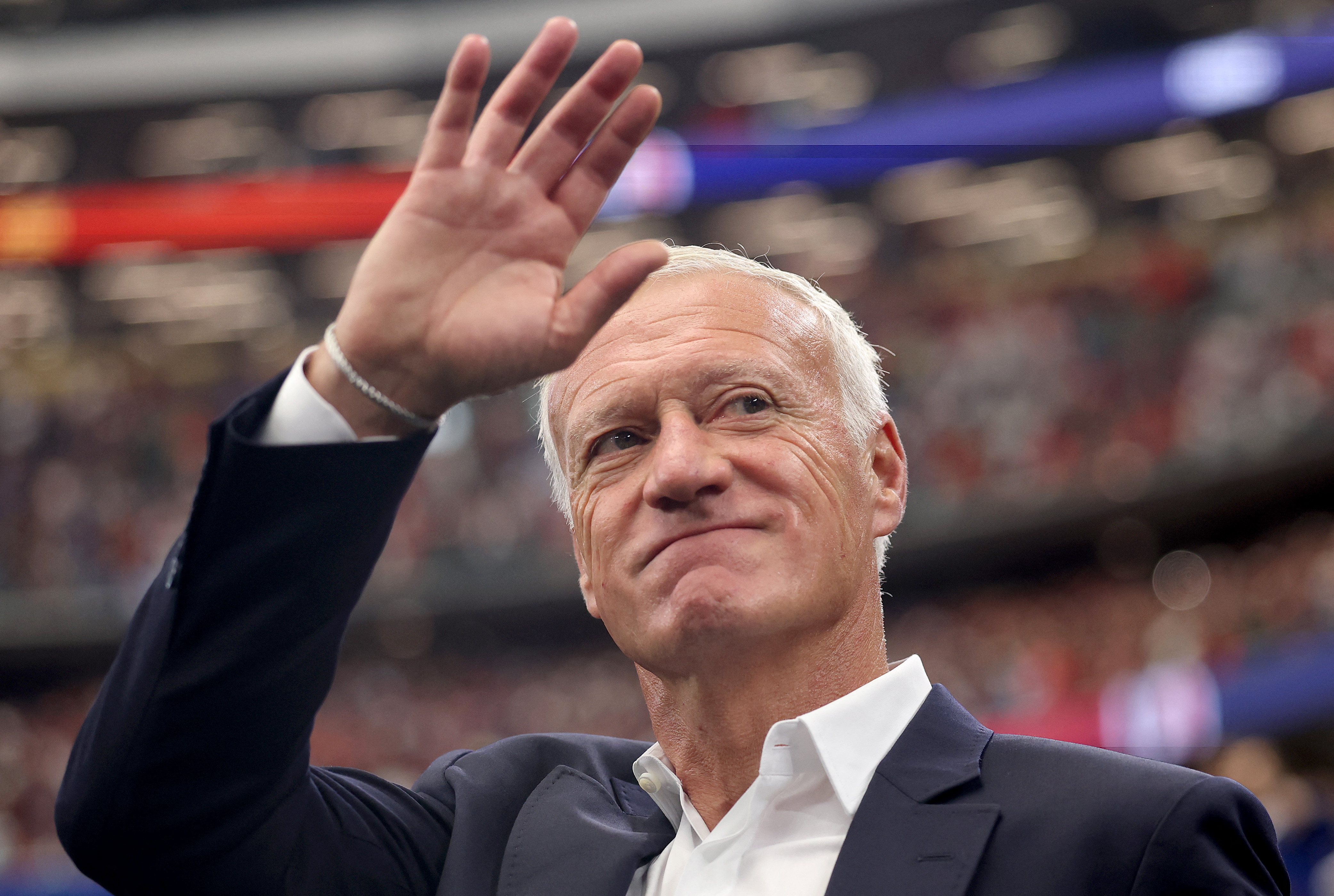 Didier Deschamps waving and smiling with fans in the background at a stadium.