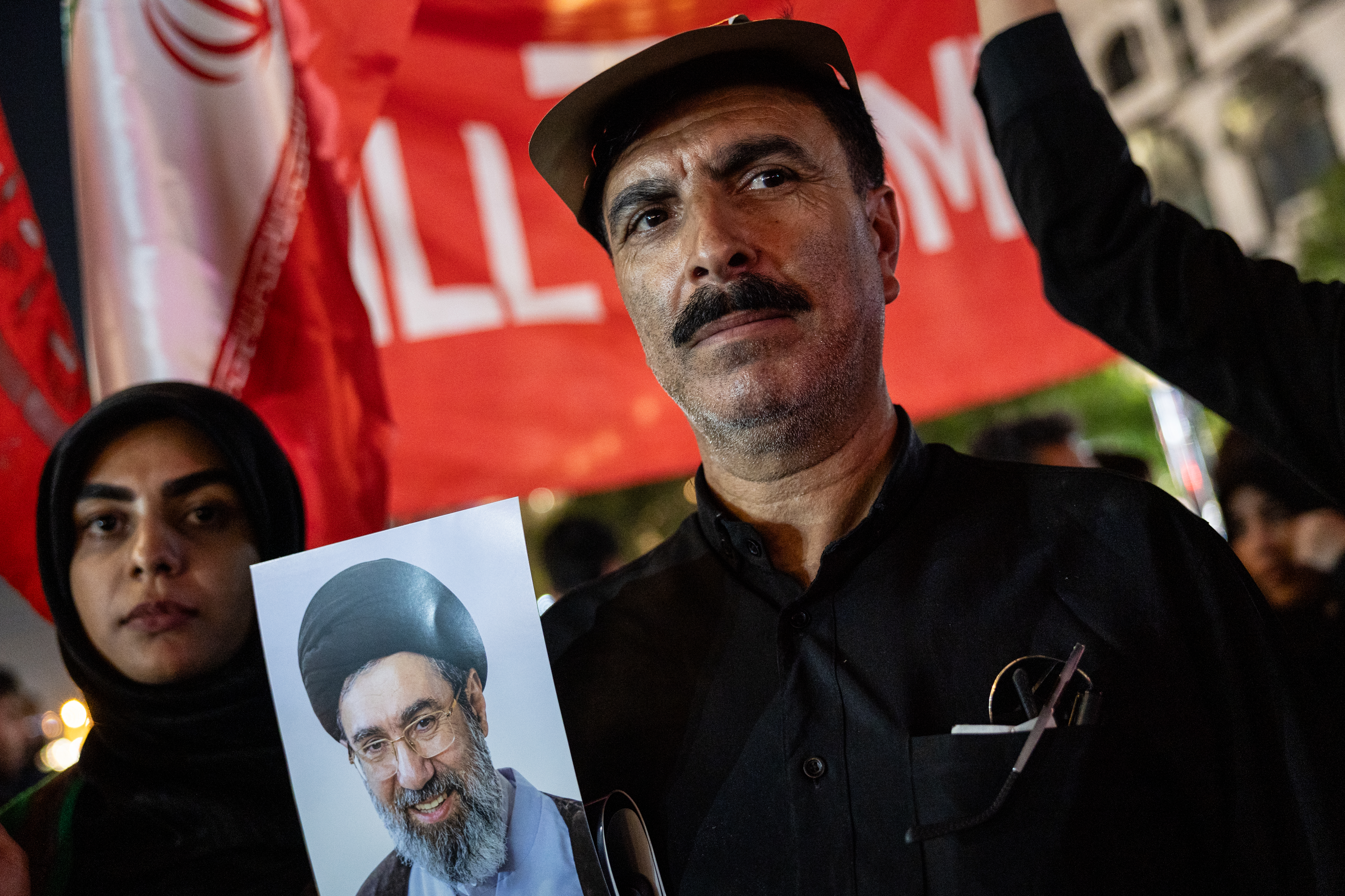 MASHHAD, IRAN - JULY 09: Mourners pose with a photo of Iran's new Supreme Leader Mojtaba Khamenei under an anti-Trump banner set up in a main thoroughfare near the Imam Reza Shrine early on July 09, 2026 in Mashhad, Iran. Thousands of people were arriving to Mashhad, Iran's spiritual capital and second largest city, to witness the arrival of slain Supreme Leader Ayatollah Ali Khamenei to be buried at the shrine, the holiest site for Shia Muslims in Iran. Khamenei and members of his family were killed on February 28 during U.S.-Israeli strikes at the beginning of the war, bringing an end to his 36-year rule over Iran. Tensions between the U.S. and Iran have escalated after the U.S. launched a new wave of strikes against Iran in response to Iranian attacks on commercial vessels transiting the Strait of Hormuz. Iran's Islamic Revolutionary Guard Corps (IRGC) said it retaliated by targeting U.S. assets in Bahrain and Kuwait, raising fears the exchange could undermine last month's agreement between Washington and Tehran to extend their ceasefire and begin negotiations to end the war. (Photo by John Moore/Getty Images)