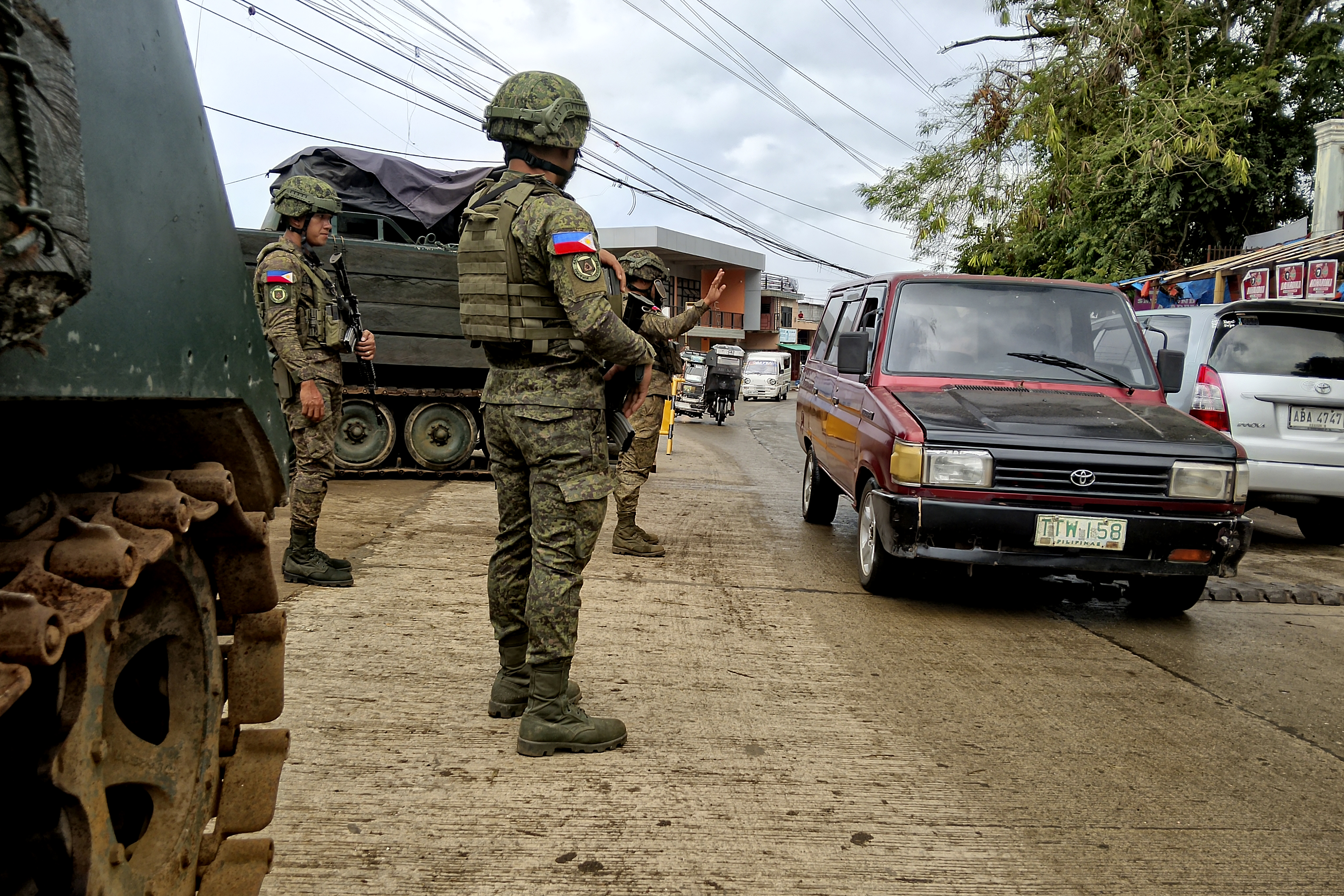Soldiers in camouflage uniforms stand at the roadside watching vehicles pass.