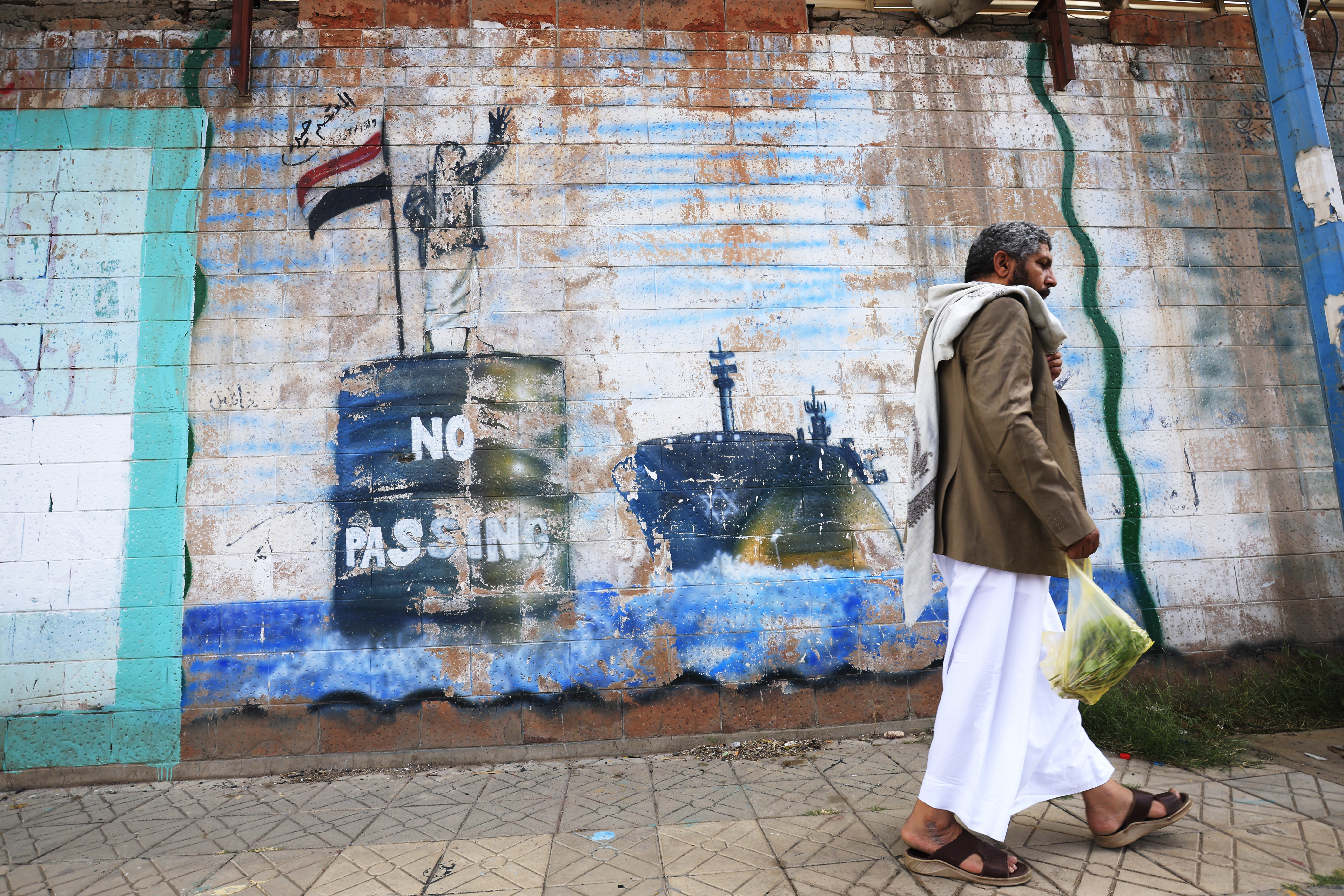 A person walks past a graffitied wall depicting a Houthi fighter stopping a ship off the coast of Yemen.