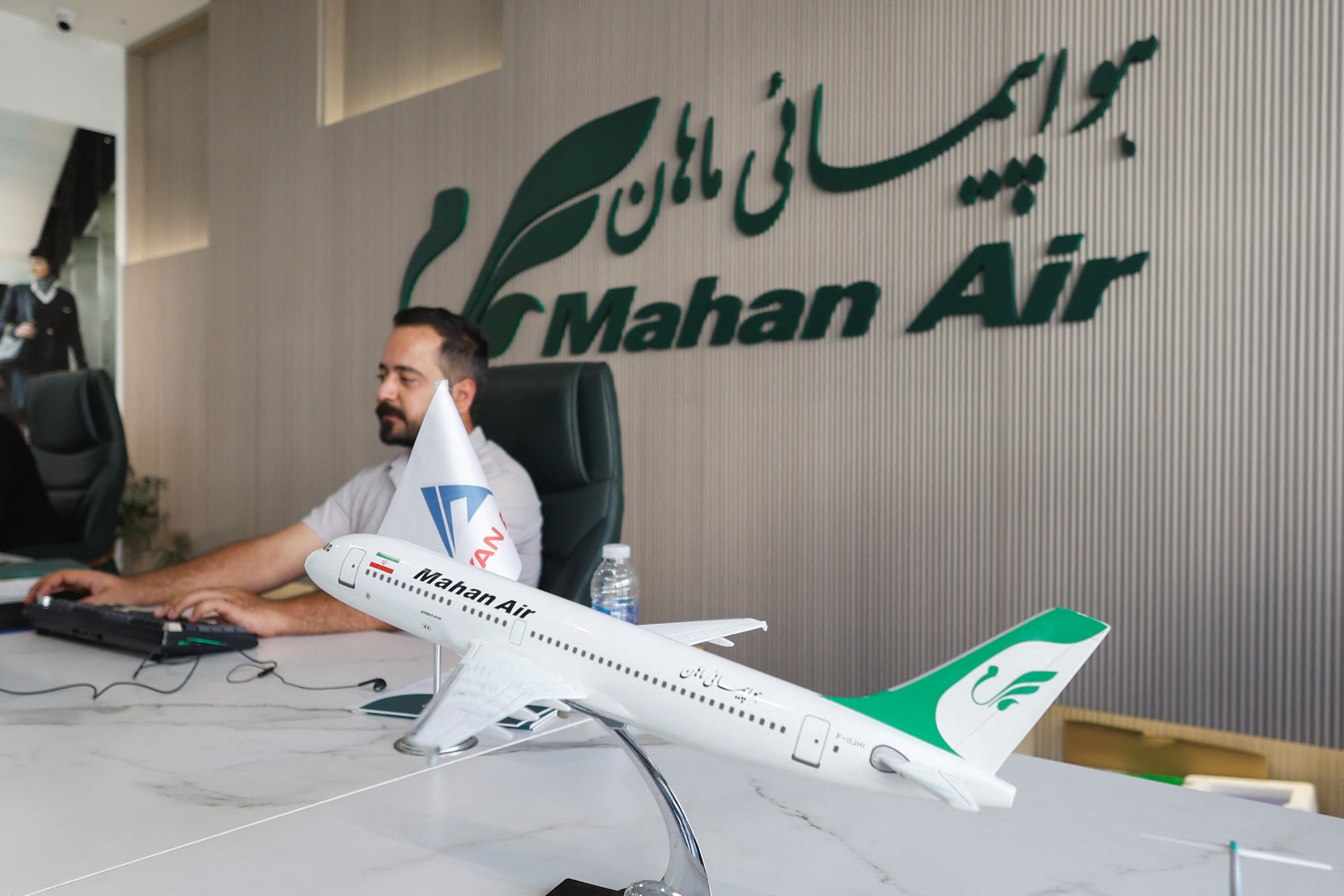 An employee works at a desk with the logo of Iran's Mahan Air behind him.